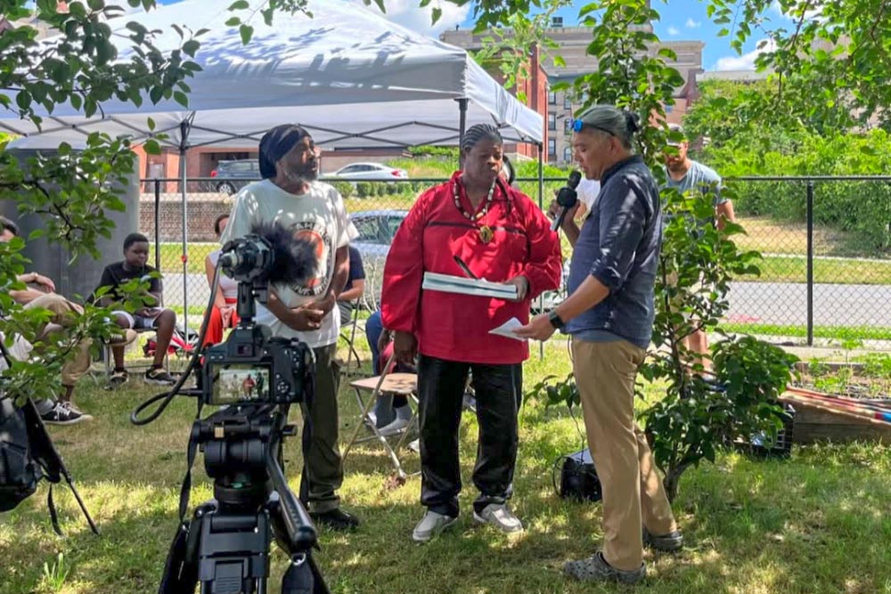 Media Coverage at a Community Garden Event Two people being interviewed on camera at an outdoor community garden event, with a pop-up tent behind them.
